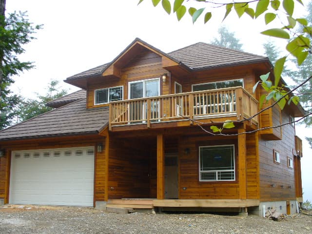 A finished two-storey home with cedar siding, a balcony and a shingle roof, trees behind it.