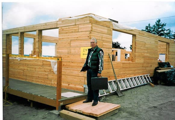 Chuck Dudley in a work jacket on a job site, standing at the deck of a square-log home under construction near the water.