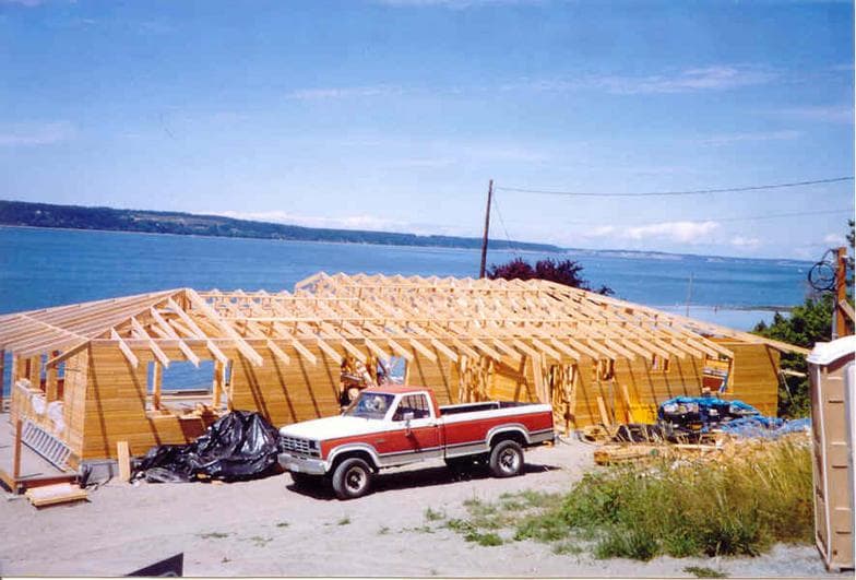 A large home under construction, its roof framing newly up, with a pickup parked in front and Puget Sound behind it.
