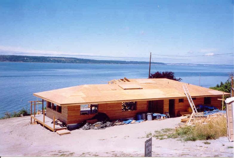 A single-storey home with its roof sheathing newly laid, overlooking the water on a clear day.